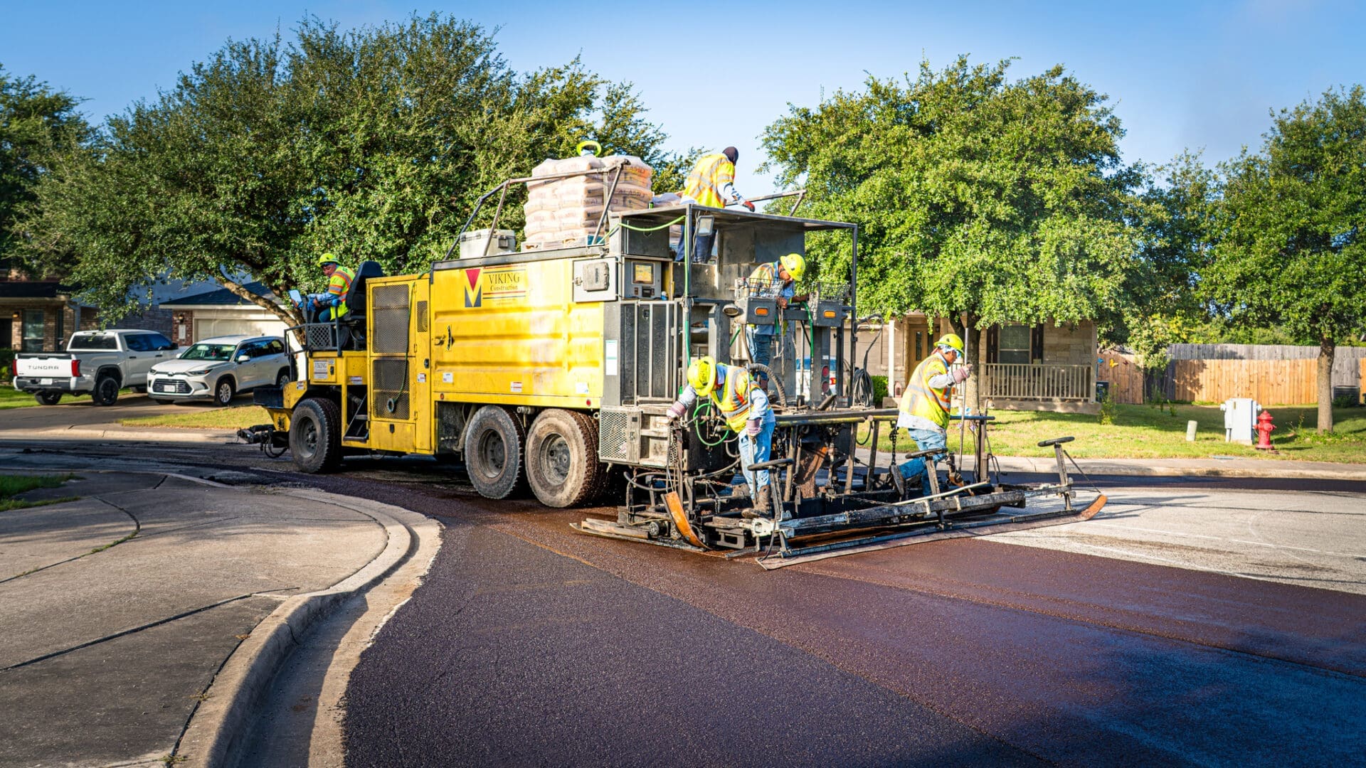 Workers operate a large yellow road paving machine, laying fresh asphalt on a residential street with trees and houses in the background on a sunny day.