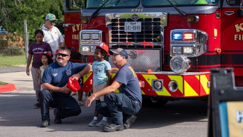 Two firefighters crouch next to a smiling child in front of a fire truck, while other children and adults stand nearby. The scene appears friendly and casual, with the fire truck’s lights visible behind them.