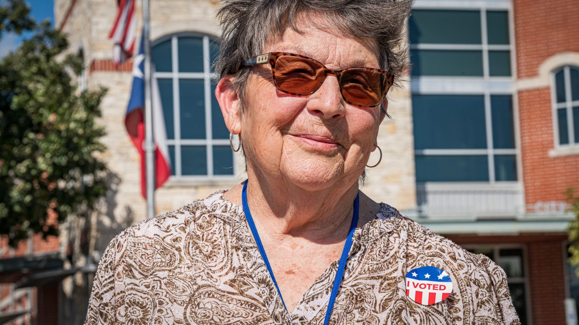 An older woman wearing sunglasses and a patterned shirt stands outside a building, smiling. She has a sticker on her chest that reads "I Voted" with stars and stripes, and U.S. flags are visible in the background.