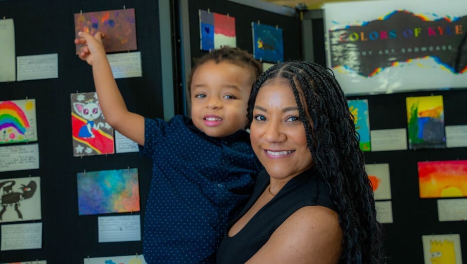 A woman holds a smiling child who is pointing at colorful artwork displayed on a wall behind them at the Tiny Art Showcase at the Kyle Public Library. Both appear happy, and the art exhibit sign reads "Colors of Kyle.