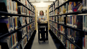Emerson pushes a book cart down an aisle between bookshelves in the Kyle Public library, surrounded by colorful books. The person wears a staff badge and appears to be shelving or organizing books.