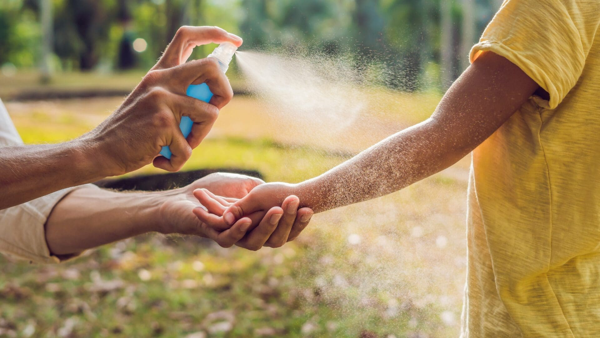 A person sprays insect repellent onto a child’s arm outdoors, with sunlight and greenery in the background.
