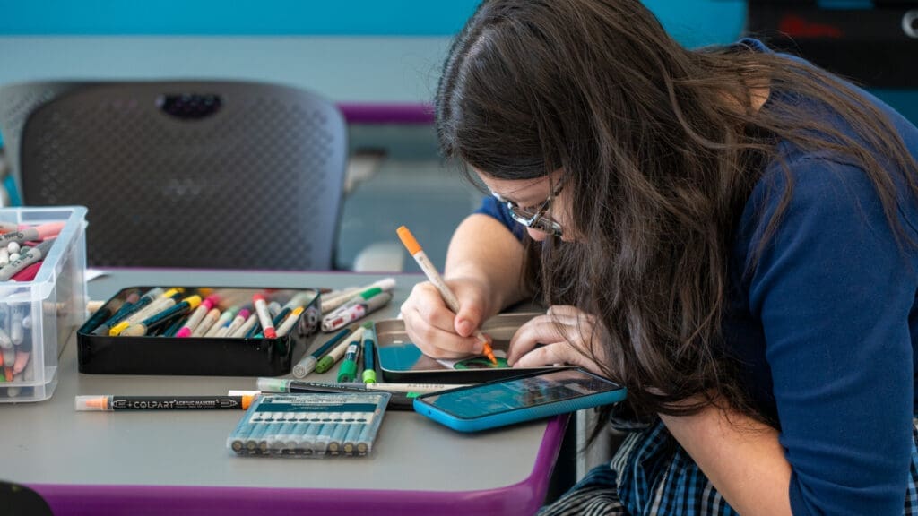 A person with long brown hair and glasses uses a tablet and marker to draw at a desk covered with markers, colored pencils, and art supplies.