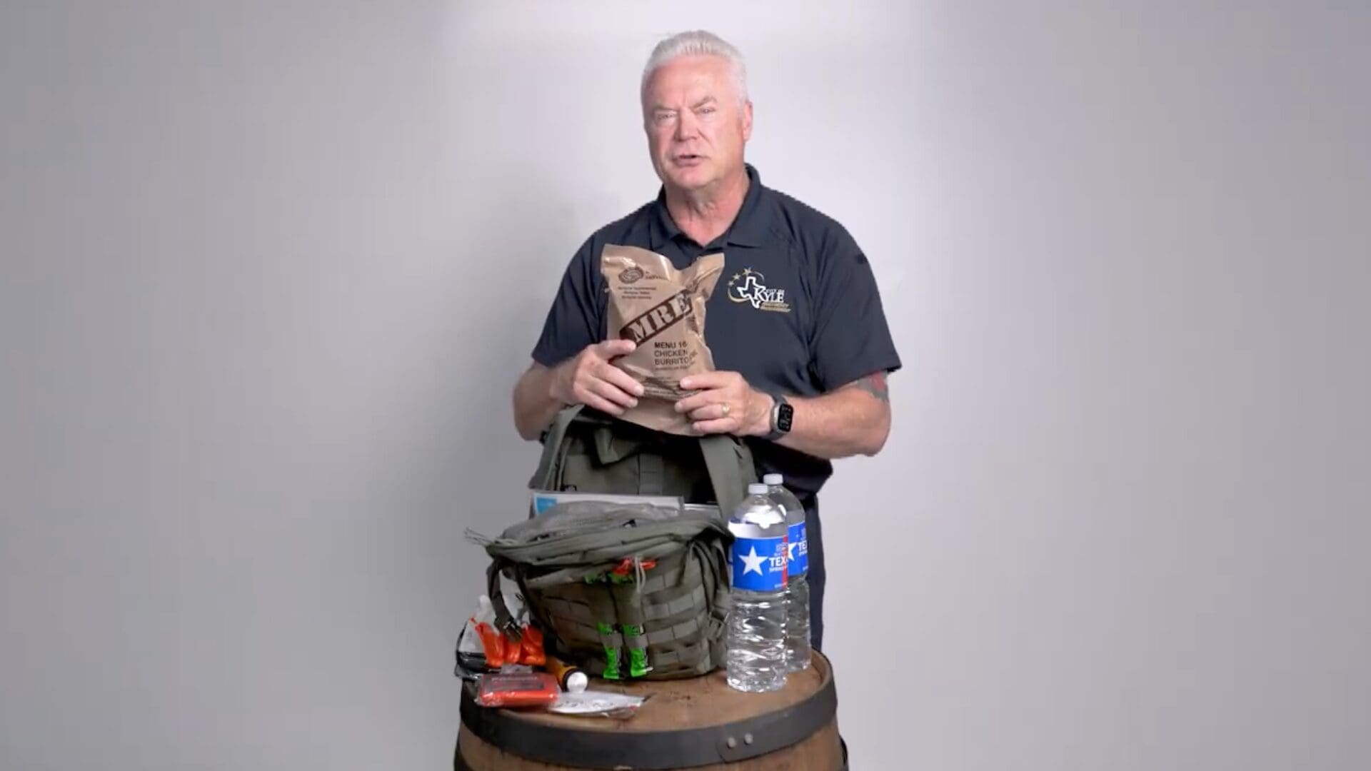 A man in a dark shirt holds a brown MRE pouch above a green bag filled with emergency supplies, including water, a flashlight, and snacks, on a wooden barrel against a plain gray background.