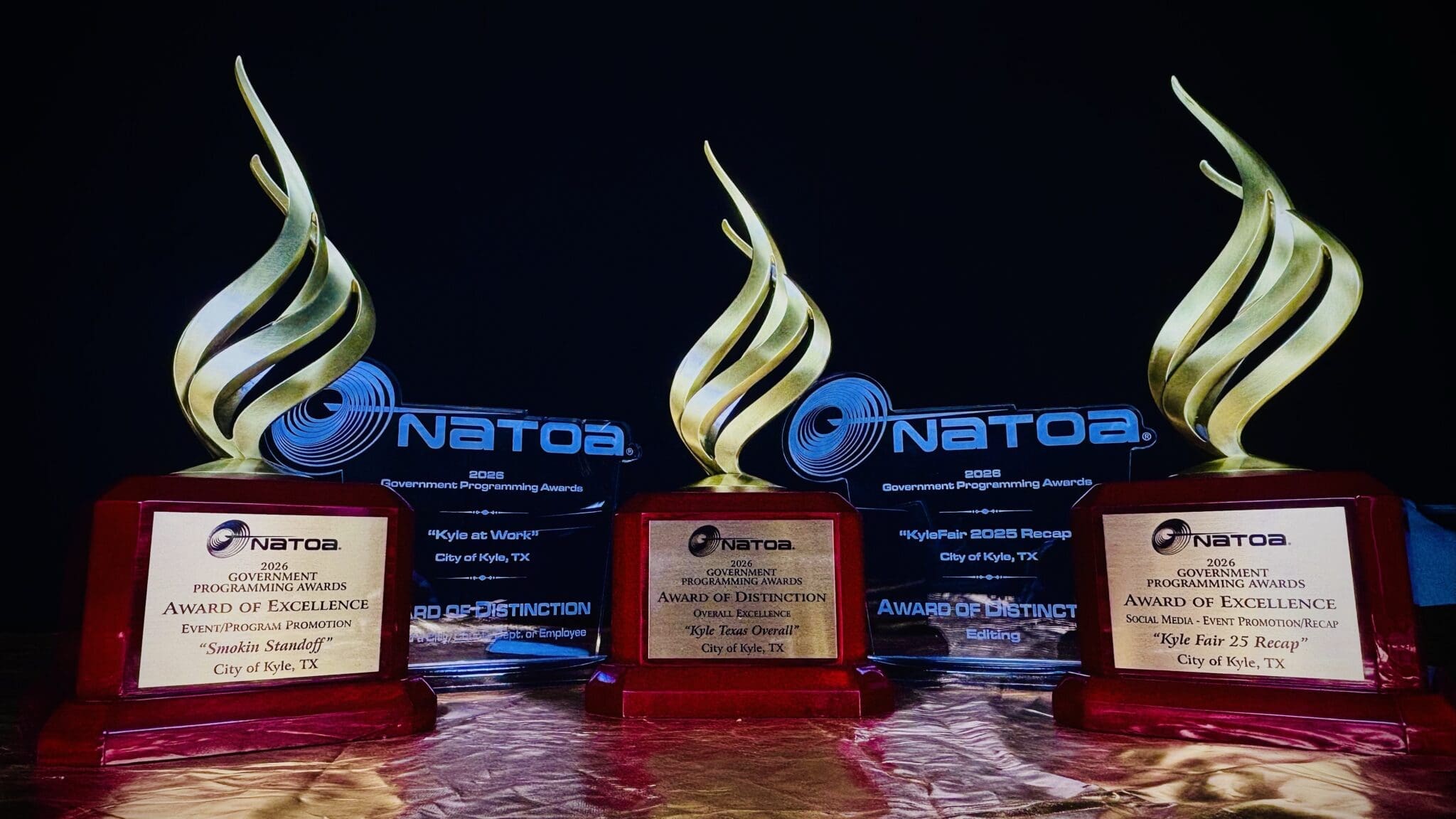 Three NATOA 2026 Government Programming Awards trophies with gold flame-shaped finials, each on a wood-tone base with engraved award details for the City of Kyle Communications Department.
