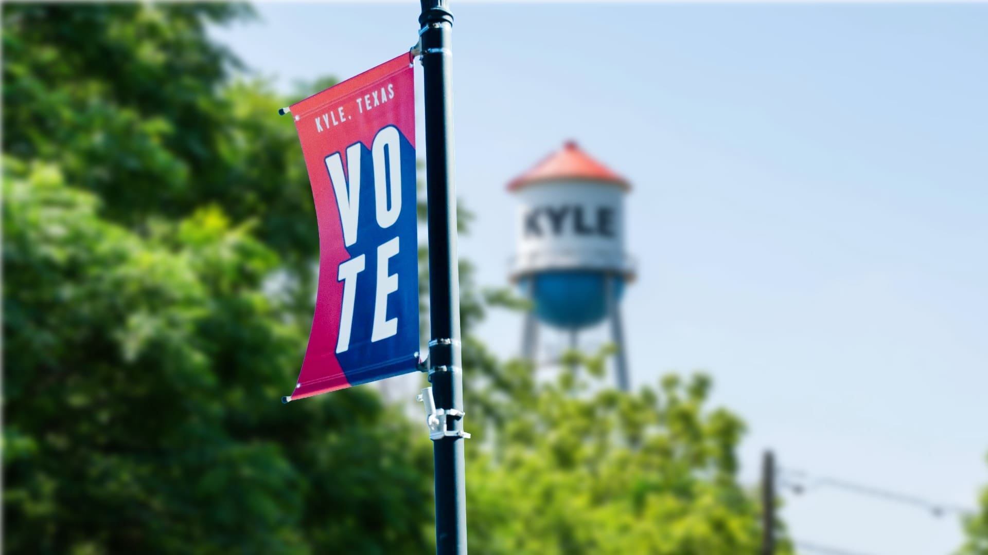 A red and blue banner on a pole reads "KYLE, TEXAS VOTE" with a water tower labeled "KYLE" and green trees blurred in the background.