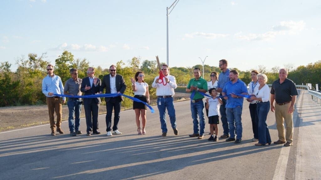 A group of people stand in a row at a ribbon-cutting ceremony on a road. One person holds large scissors, cutting a blue ribbon, while others look on and smile under a clear sky.
