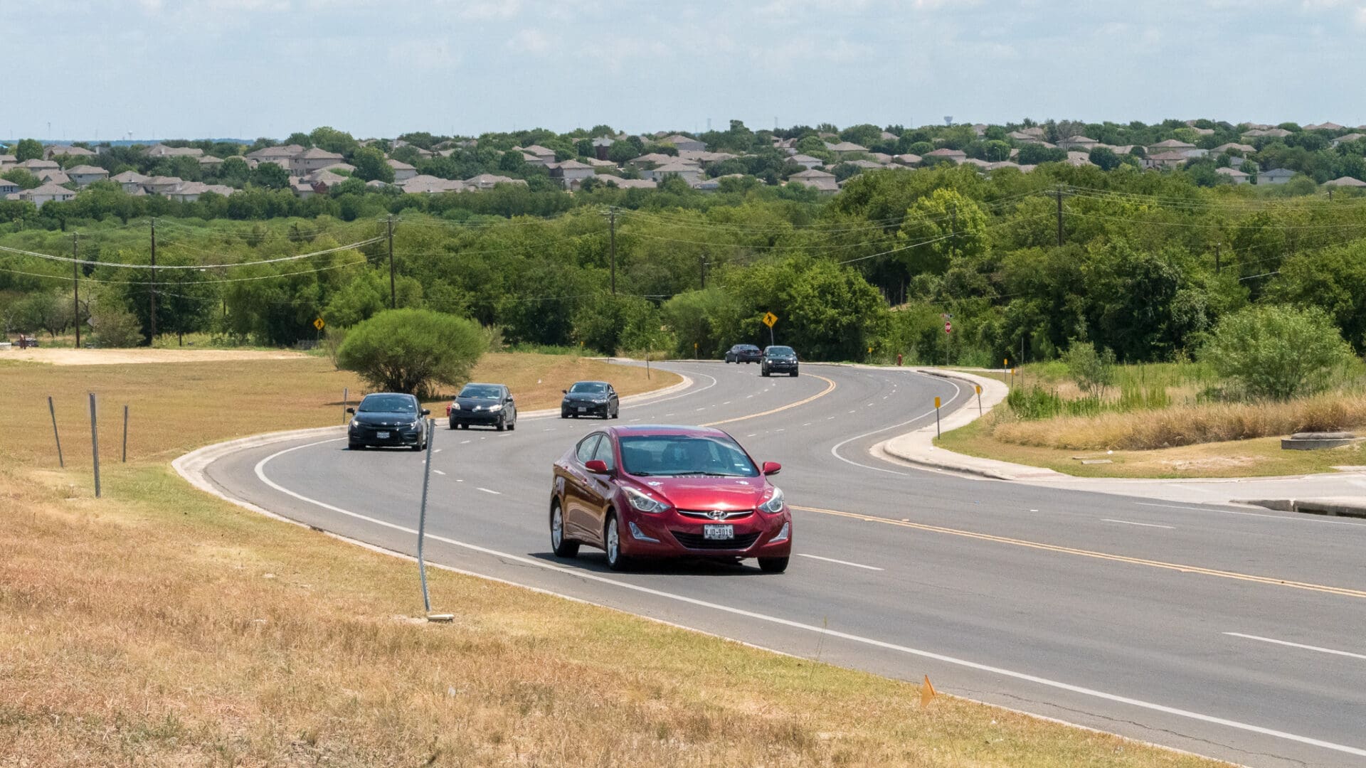 A red car leads several vehicles driving on a curving, two-lane road bordered by grassy fields and trees, with houses visible in the distance under a partly cloudy sky.