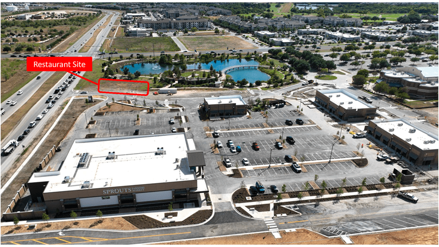 Aerial view of a commercial area with a grocery store (Sprouts Farmers Market) in the foreground, parking lots, nearby buildings, a pond, and a red label marking a "Restaurant Site" on an empty lot near a busy road.