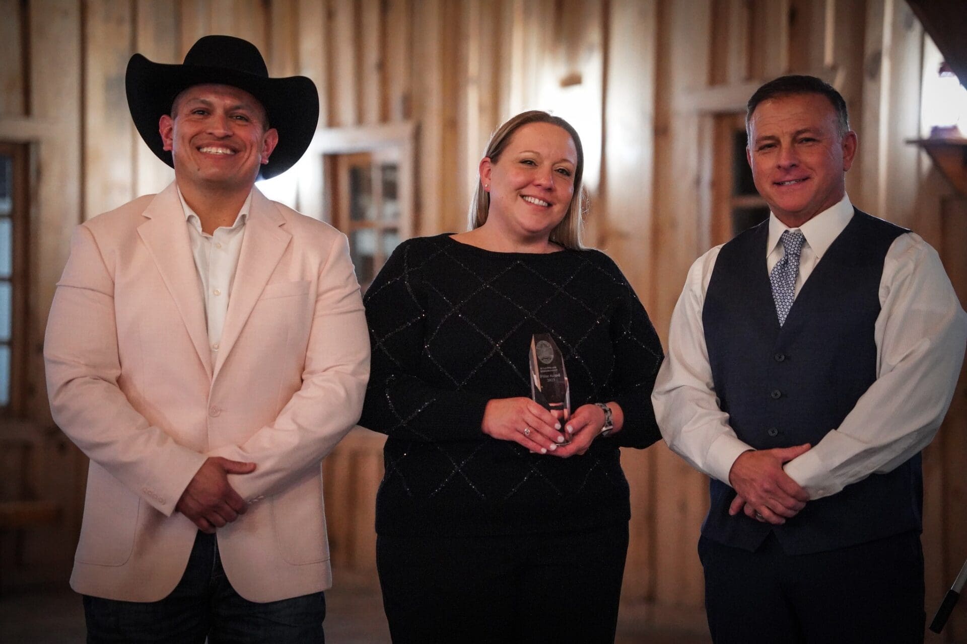Three people stand indoors in front of a wooden wall; the man on the left wears a light cowboy hat and jacket, the woman in the middle holds an award, and the man on the right wears a suit vest and tie. All are smiling.