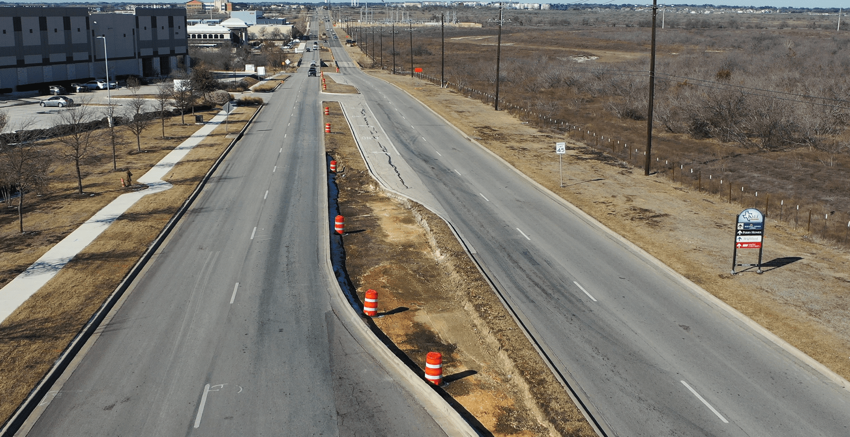 Aerial view of a divided road under construction, with orange traffic barrels along the median. One side of the road is bordered by a sidewalk and buildings; the other by a dry, undeveloped field.