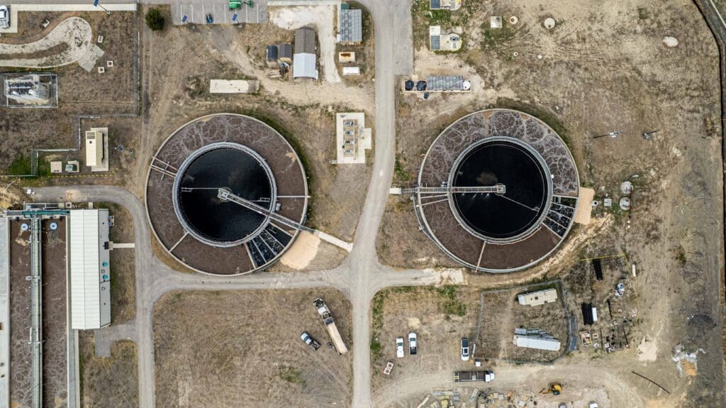 Aerial view of a wastewater treatment plant showing two large, circular settling tanks surrounded by roads, small buildings, parked vehicles, and dry, brown land.