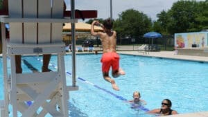 A boy in orange swim trunks jumps into an outdoor swimming pool near a lifeguard chair, while two people smile and watch him from the water on a sunny day.