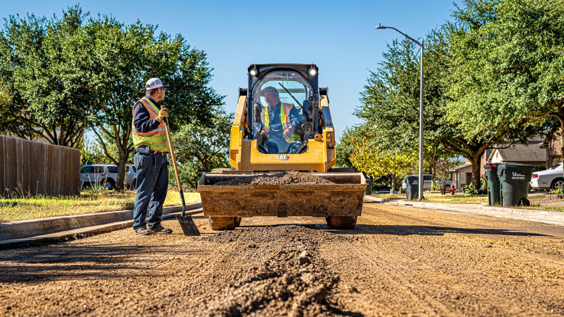 Two City of Kyle PublicWorks employees in safety vests repair a street; one operates a small bulldozer while the other stands nearby with a shovel. The street is covered in dirt, and trees and houses line the background.