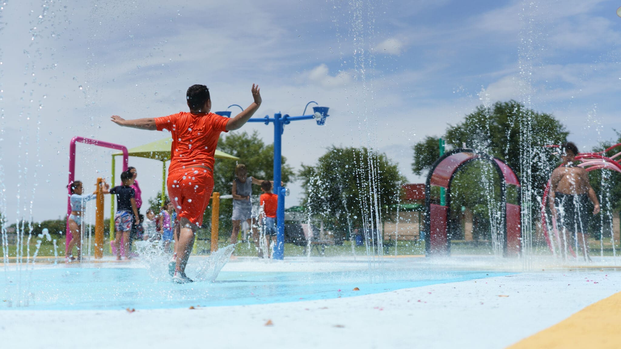 A child in an orange outfit runs through water jets at a splash pad, with other children playing in the background under a partly cloudy sky.